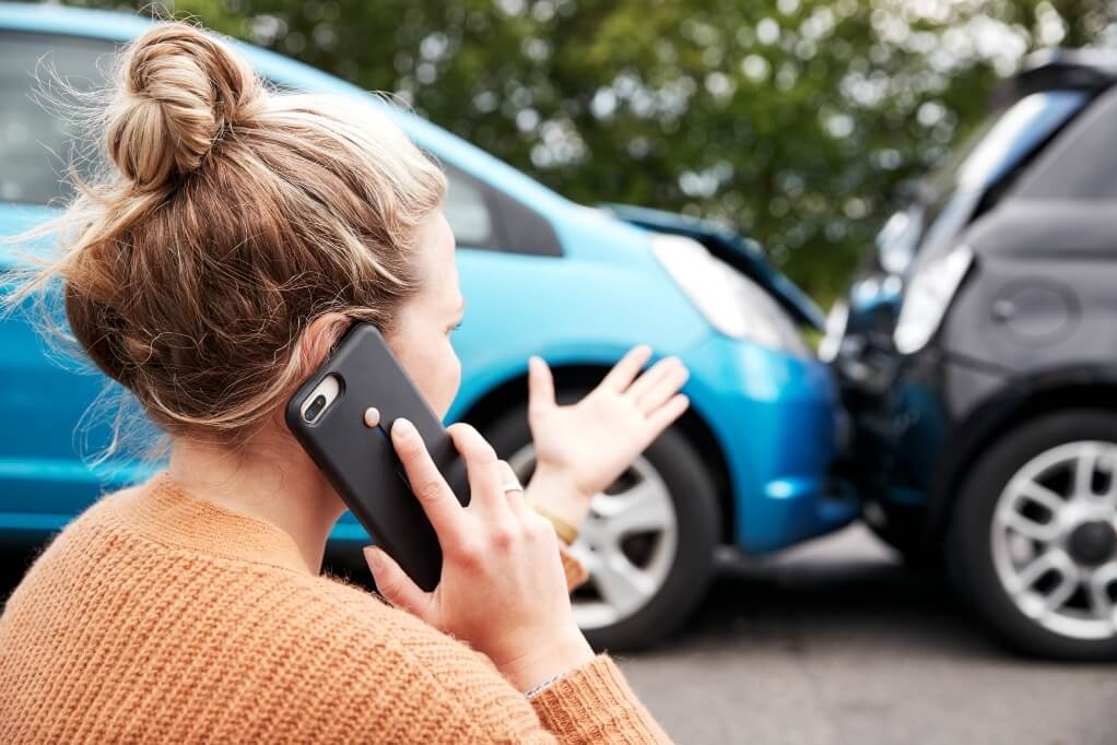 a concerned woman in front of a damaged car, talking on her cell phone