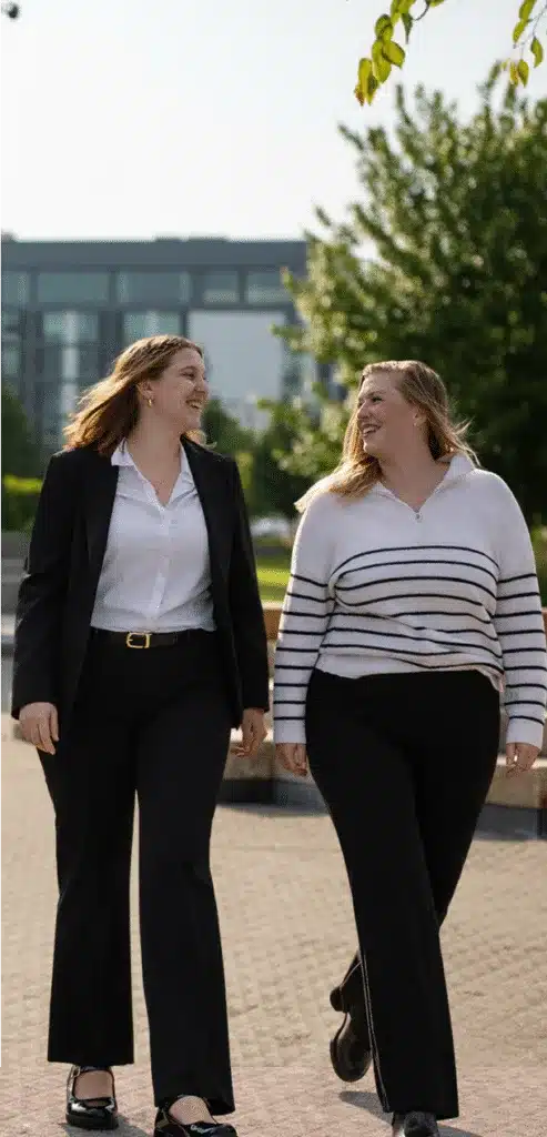 two smiling professional women walk outdoors in business attire chatting beneath leafy trees
